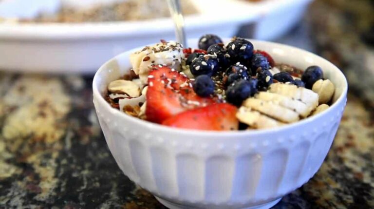 picture of a smoothie with granola, blueberries, strawberries, and bananas in a white bowl on a counter from "Acai Smoothie Bowl" by Green Smoothie Girl