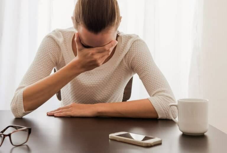 Photo of woman holding her forehead in her hand and cellphone on the table from "28 Signs That You Are Electrosensitive" by Green Smoothie Girl