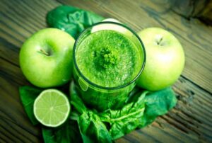 Photograph of a green smoothie in a glass surrounded by apples, limes, and leafy greens, from "9 Ways to Get Kids to Drink Healthy Green Smoothies"