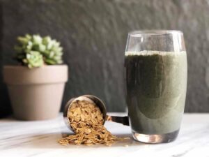 Photo of green smoothie in glass with measuring cup spilling out oats with plant in background from "Oatmeal Breakfast Smoothie" recipe by Green Smoothie Girl