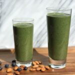 Photo of two green smoothies in a big and small glass with blueberries and almonds scattered on a wood surface with marble in the background from "Super Hulk Green Smoothie" recipe by Green Smoothie Girl