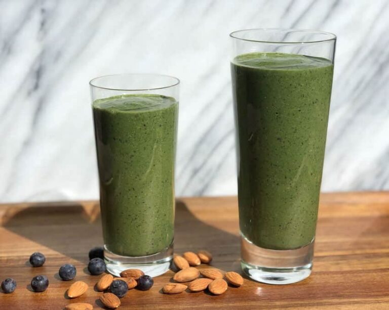 Photo of two green smoothies in a big and small glass with blueberries and almonds scattered on a wood surface with marble in the background from "Super Hulk Green Smoothie" recipe by Green Smoothie Girl