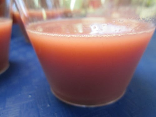 Photograph of a pink drink in a small transparent cup, sitting on a blue tablecloth.