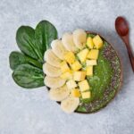 a green smoothie in a wooden bowl with sprinkles of chia seeds and chucks of banana and mango next to spinach leaves and a wooden spoon on a gray slate background from Green Smoothie Girl's "smoothie bowl"