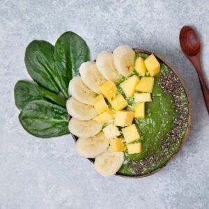 a green smoothie in a wooden bowl with sprinkles of chia seeds and chucks of banana and mango next to spinach leaves and a wooden spoon on a gray slate background from Green Smoothie Girl's "smoothie bowl"