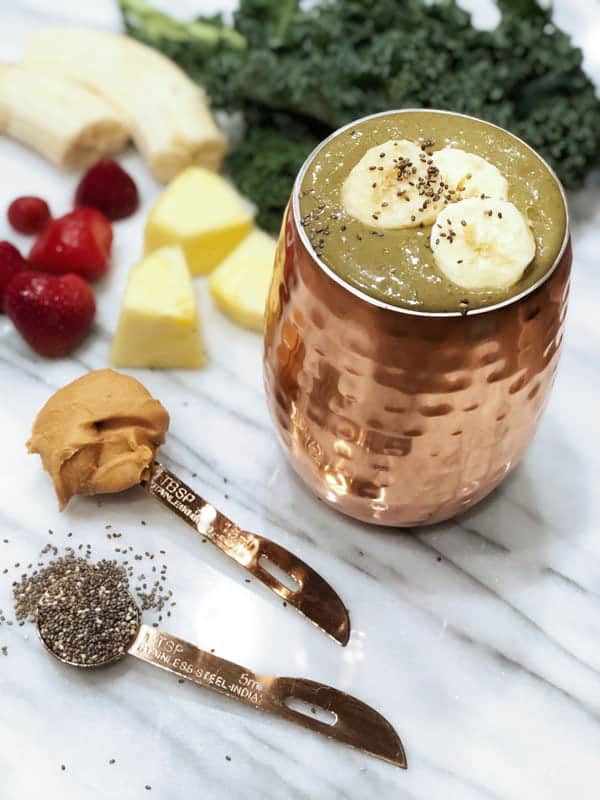 Photo of green smoothie in copper cup with kale, banana, strawberries, pineapple, banana, chia seeds, peanut butter surrounding from "Banana Split Smoothie" recipe by Green Smoothie Girl