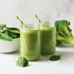 A photo of two green smoothies with green and white straws next to a broccoli floret and a white bowl of spinach with a white background from Green Smoothie Girl's "Broccoli Blitz Smoothie"