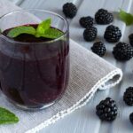 Photo of purple smoothie in glass on grey table runner with surrounding blackberries from "Mustard Greens Mambo" recipe by Green Smoothie Girl