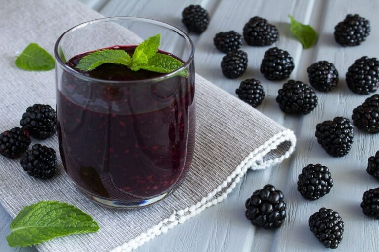 Photo of purple smoothie in glass on grey table runner with surrounding blackberries from "Mustard Greens Mambo" recipe by Green Smoothie Girl