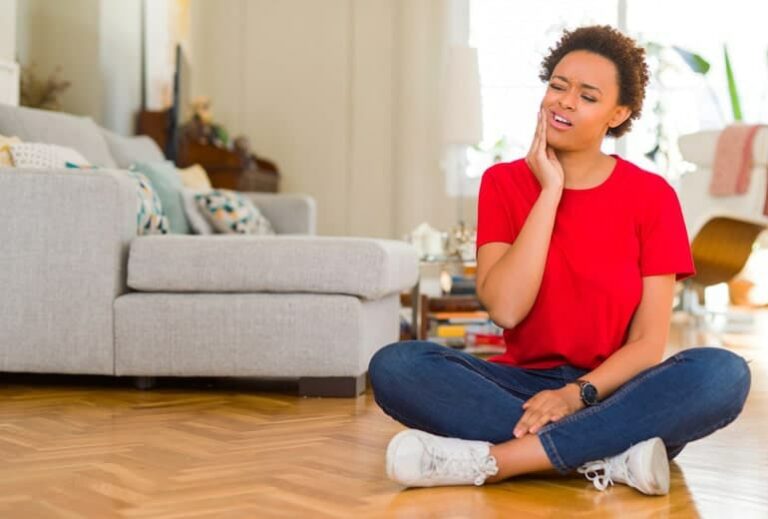 Photo of young black woman sitting on floor holding jaw in pain from "What Really Causes Cavities, and What Can You Do to Prevent Them?" by Green Smoothie Girl