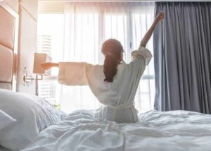 Photo of young Asian woman sitting on bed towards sun stretching from "14 Ways I Optimize My Health And Energy Every Day" by Green Smoothie Girl