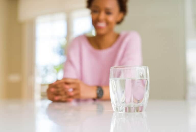 Photo of glass of clean water on table with African American woman smiling in background from "3-Day Water Fasting vs. 3-Day Modified Fasting" by Green Smoothie Girl