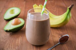 Photo of chocolate brown Muddy Buddy green smoothie with striped straw inserted and bananas and avocados surrounding it from "11 Green Smoothie Recipes for Fatty Liver" by Green Smoothie Girl