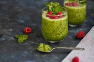 Photo of Dandelion Doodleberry Juice green smoothie recipe with green smoothie in glasses with raspberries on top and surrounding from "11 Green Smoothie Recipes for Fatty Liver" by Green Smoothie Girl