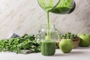 Photo of 4-3-2-1 green smoothie recipe being poured from blender into glass with apples and parsley surrounding from "11 Green Smoothie Recipes for Fatty Liver" by Green Smoothie Girl