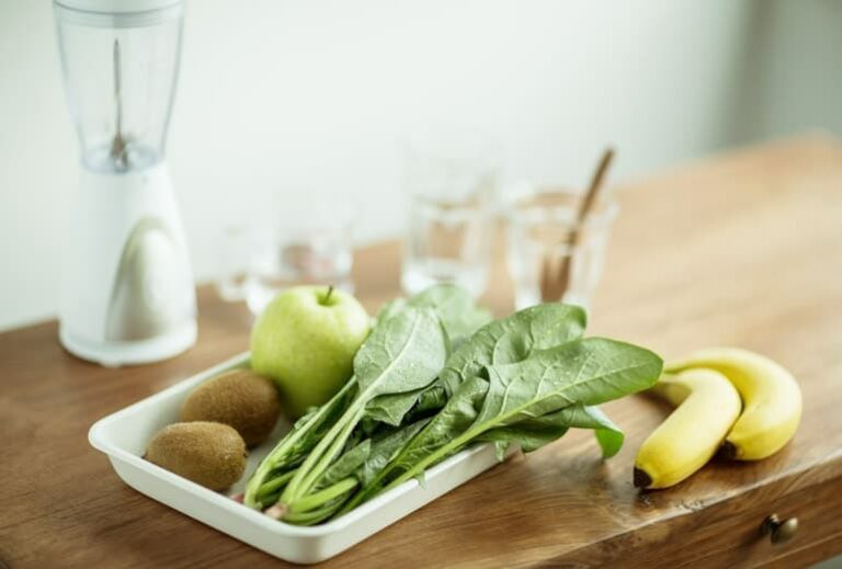 Photo of spinach, apple, and kiwis in tray on wooden table with blender and glass cups in background from "15 Tasty Meal Replacement Smoothie Recipes To Fill You Up & Keep You Going" blog post by Green Smoothie Girl