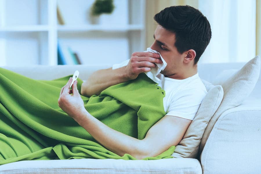 A sick man lays on the couch under a green blanket.