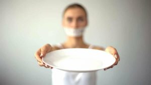 Photo of skinny woman with taped mouth holding up an empty plate from "The Truth About The OMAD (One Meal a Day) Diet" by Green Smoothie Girl