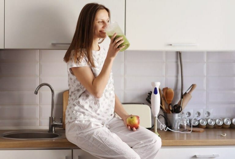 A woman drinking a green smoothie while sitting on the kitchen counter from Green Smoothie Girl