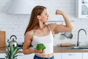 A strong woman flexing her muscles while holding a broccoli in a kitchen.