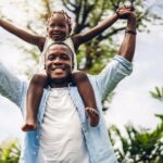 A smiling man holding his smiling daughter on his shoulders outside