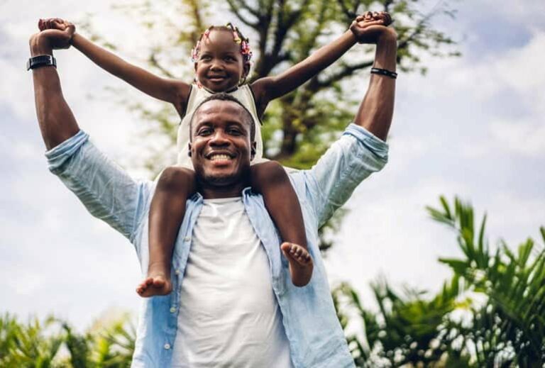 A smiling man holding his smiling daughter on his shoulders outside