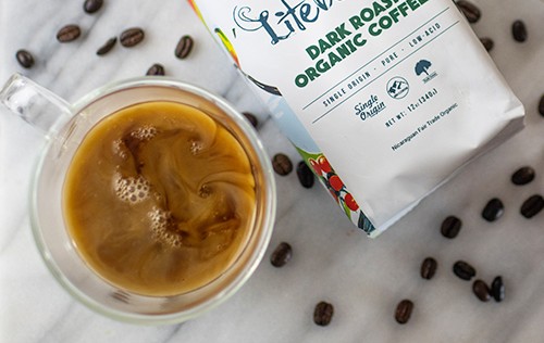 A cup of coffee with a package of LifeBoost coffee next to the cup. There are coffee beans on the table around the cup and package from Green Smoothie Girl's "Easy Bone Broth Coffee Latte"