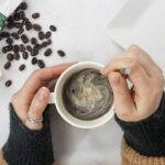 a woman's hands stirring coffee in a white mug . There are coffee beans on the table from Green Smoothie Girl's "Immune-Boosting Bone Broth Coffee Elixir"