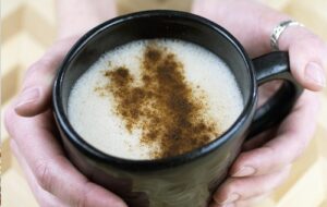 A woman holding a black mug of white hot chocolate with spices on top from Green Smoothie Girl's "Sweet and Spicy Vanilla Protein Elixir"