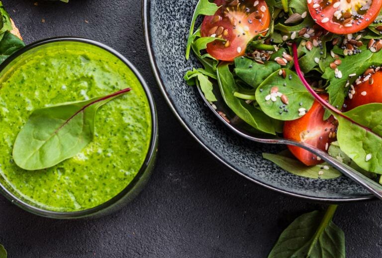 a salad in the upper right corner and a green smoothie with a spinach leaf over top of it with a gray background from Green Smoothie Girl's "Blended Salad"