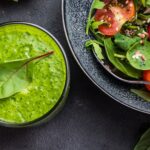 A green salad in the upper right corner next to a green smoothie with a leaf of spinach in it with a gray background from Green Smoothie Girl's "Blended Salad"