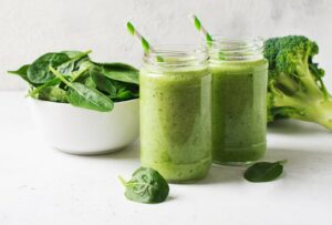 A photo of two green smoothies with green and white straws next to a broccoli floret and a white bowl of spinach with a white background.