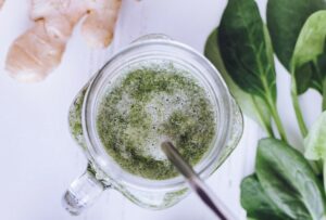 Photo of a green smoothie in a clear jar mug with a metal straw next to several spinach leaves and a ginger root on a white background.