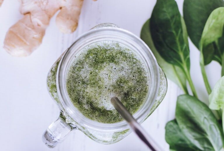 Photo of a green smoothie in a clear jar mug with a metal straw next to several spinach leaves and a ginger root on a white background from Green Smoothie Girl's "Chai Green Smoothie"