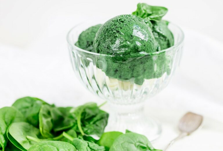 Several scoops of green ice cream in a clear bowl next to a pile of spinach from Green Smoothie Girl's "Green Ice Cream"