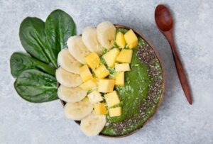 A green smoothie in a wooden bowl with chunks of mango and banana and a sprinkling of chia seeds. There are spinach leaves and a wooden spoon next to the bowl on a gray slate background.