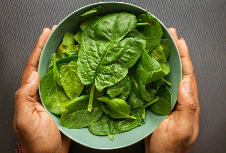 hands holding a green bowl of spinach with a dark gray background from Green Smoothie Girl's "9 of the Best Spinach Smoothies — Easy, Delicious, and Healthy"