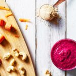 photo of a hot pink breakfast smoothie and a cup of protein powder next to a cutting board with various chopped vegetables and nuts from Green Smoothie Girl's "11 Delicious High-Protein Smoothies to Fill You Up for Hours"