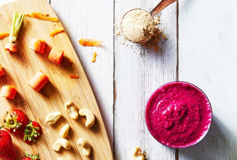 photo of a hot pink breakfast smoothie and a cup of protein powder next to a cutting board with various chopped vegetables and nuts from Green Smoothie Girl's "11 Delicious High-Protein Smoothies to Fill You Up for Hours"