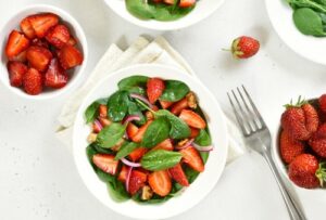 image of a bowl of spinach and strawberries with several bowls of individual strawberries surrounding it from Green Smoothie Girl's "spinach strawberry salad" recipe
