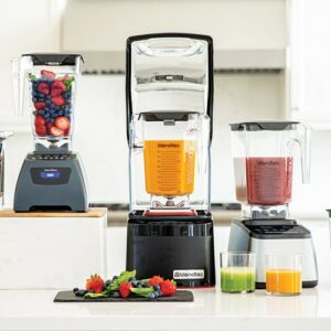 3 different style of Blendtec blenders. 1 is full of fruit, 1 is full of an orange smoothie, 1 is full of a pink smoothie. a cutting board of fruit and 2 glasses of smoothie sit in front of the blenders