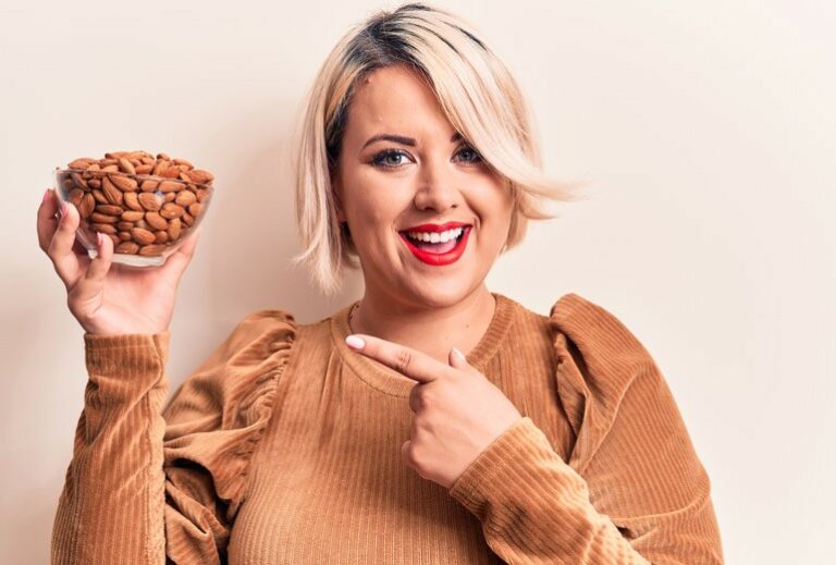 A woman with short dyed blonde hair is smiling and pointing at a clear bowl of almonds from Green Smoothie Girl's "Surprising Health Benefits of Almonds for Immunity" recipe