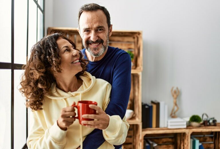 a woman holding a mug smiles up at her husband who is hugging her from behind