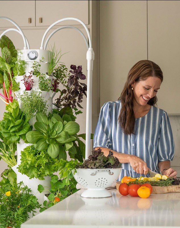 Woman chopping vegetables on the counter in her kitchen. She is standing next to a Tower Garden full of leafy green veggies